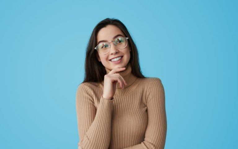 A smiling woman with long dark hair and glasses in a beige sweater poses confidently.