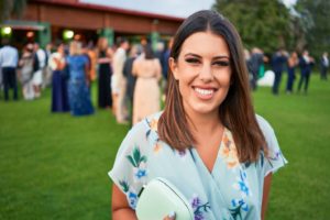 A woman in a floral dress smiles warmly at an outdoor event.