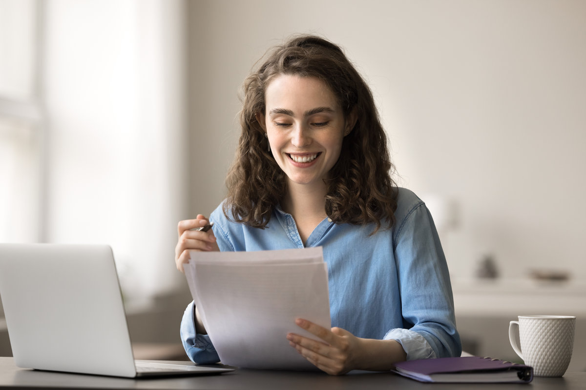 Smiling attractive student woman sit at desk with laptop, read essay, makes correction, feels satisfied with work done, prepare for session, working on diploma, filling form, doing examination test